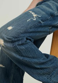 Modre jeans hlače s sproščenim krojem, ki vključujejo obrabljene obliže in strgane detajle na kolenih, kar prikazuje raznoliko teksturo.