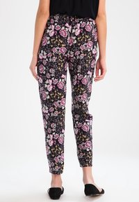 Une personne portant un pantalon noir avec des motifs floraux roses et jaunes et des chaussures plates noires, de dos par rapport au spectateur.