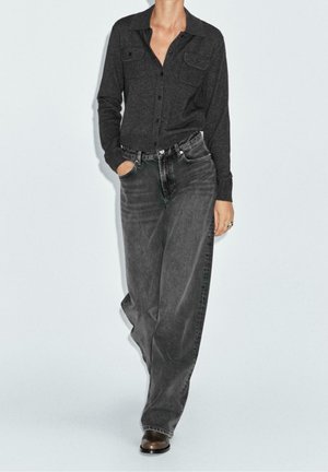 Kvinde iført en mørkegrå skjorte med knapper og løstsiddende falmede sorte jeans, stående med den ene hånd i lommen mod en lys baggrund.