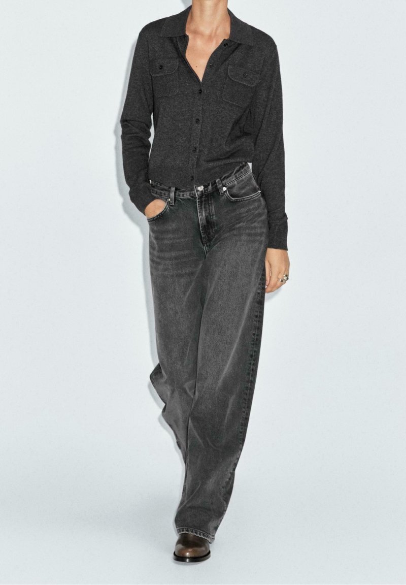Femme portant une chemise boutonnée gris foncé et un jean noir délavé ample, debout avec une main dans la poche sur un fond clair.