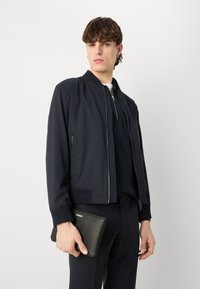 Morska bomberska jakna z rebrastimi manšetami, zadrgo spredaj in stranskimi žepi. Model drži črn usnjen clutch, na katerem je napis blagovne znamke "HUGO."