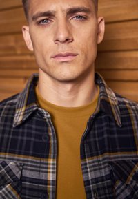 Károvaná košile s knoflíky v šedé, černé a žluté barvě přes hořčicové tričko. Detailní pohled ukazuje límec a texturu látky.