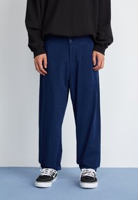 Pantalones azul marino de corte relajado, con cierre de botón frontal, bolsillos laterales y tejido de textura suave. Se llevan con zapatillas negras y blancas.