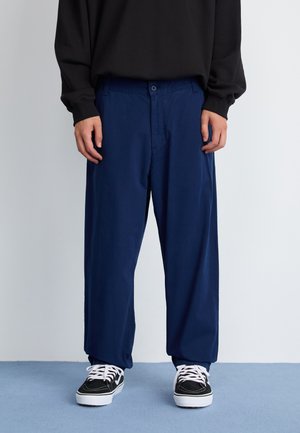 Navy blue trousers with a relaxed fit, featuring a front button closure, side pockets, and smooth textured fabric. Worn with black and white sneakers.