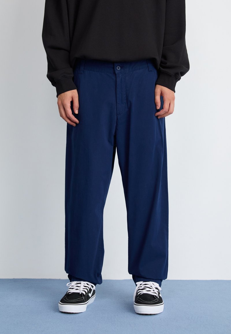 Pantalones azul marino de corte relajado, con cierre de botón frontal, bolsillos laterales y tejido de textura suave. Se llevan con zapatillas negras y blancas.