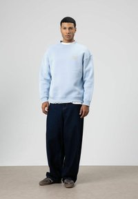 Sweat-shirt bleu clair avec un col rond et une coupe ample, associé à un jean en denim foncé. Des chaussures décontractées complètent la tenue.