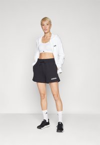 Hvit hettejakke med glidelås, svarte treningsshorts og svarte sneakers med hvite detaljer. Sportstopp synlig under hettejakken. Logo vist på klærne.