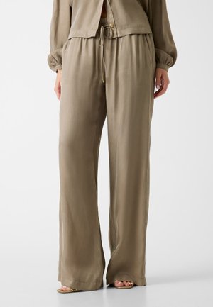 Donna che indossa pantaloni beige ampi e larghi con vita elastica e coulisse, abbinati a sandali open-toe su uno sfondo chiaro.