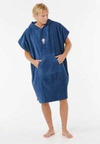 Man in een marineblauw capuchon poncho met voorzak die blootsvoets staat en zijwaarts kijkt tegen een witte achtergrond.