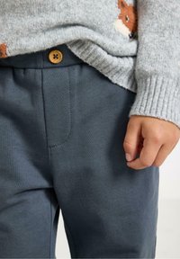 Kind in een grijze gebreide trui met vosmotief en blauwe broek met zichtbare houten knop, met de rechterhand ontspannen aan de zijde.