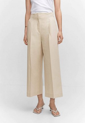 Mujer que lleva pantalones beige de pierna ancha y cortos, top blanco sin mangas y sandalias de tacón gris claro, de pie frente a un fondo sencillo.