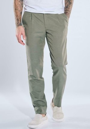 Man wearing olive green pants with front pleats and beige slip-on shoes standing against a plain background.