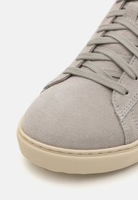 Gray suede sneaker with a rounded toe, flat beige rubber sole, and matching laces. Features subtle stitching and textured surface.