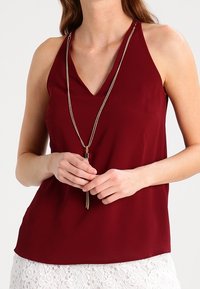 Bordowa bezrękawnikowa bluzka z dekoltem w kształcie litery V, gładka w dotyku, z cienkim metalowym naszyjnikiem z akcentem w postaci frędzla.