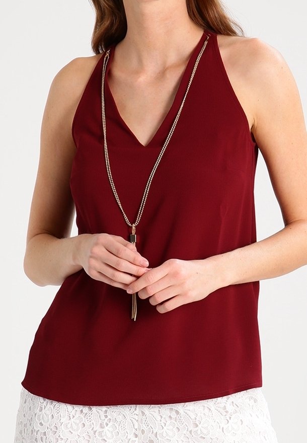 Bordowa bezrękawnikowa bluzka z dekoltem w kształcie litery V, gładka w dotyku, z cienkim metalowym naszyjnikiem z akcentem w postaci frędzla.