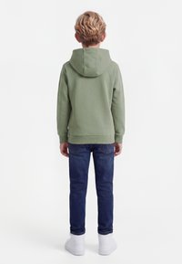 Dziecko z krótkimi blond włosami, ubrane w zieloną bluzę z kapturem, ciemnoniebieskie dżinsy i białe tenisówki, zwrócone tyłem na białym tle.