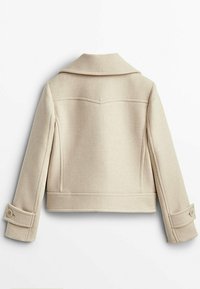 Beżowa kurtka z wełny z klapami, zapinana na guziki przy mankietach oraz z delikatnymi szwami na ramionach. Design sięgający kostek.