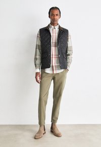 Colete preto acolchoado com fecho de botões à frente, sobre uma camisa xadrez em cinza, branco e vermelho, combinado com calças verde-oliva e botas bege.