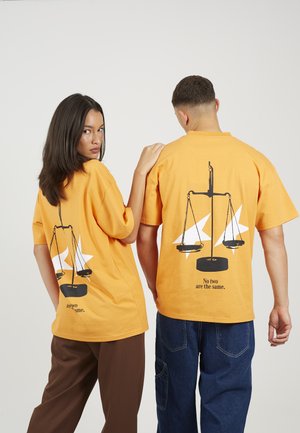 Twee mensen dragen bijpassende oranje t-shirts met een zwart schubbenpatroon en de tekst "Geen twee zijn hetzelfde", waarvan één met de voorkant naar voren en de ander met de achterkant naar voren.