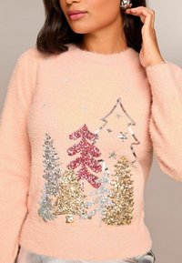 Rozā pūkaina džemperis ar izšūtu dizainu, kurā ir sekojošas sequined koki sarkanā, sudraba un zelta krāsā, papildināti ar mirdzošām zvaigznēm.