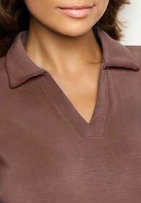 Primer plano de una persona con una camiseta marrón de cuello en V con un cuello, mostrando el cuello, la barbilla y parte del cabello rizado.