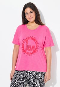 T-shirt in cotone rosa con un design grafico rotondo rosso. Maniche corte e vestibilità rilassata, abbinata a pantaloni a fantasia bianca e nera.