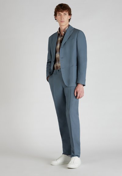 Abito blu chiaro con vestibilità su misura, abbinato a una camicia a quadri. Le caratteristiche del design includono un risvolto a punta e una tasca sul petto singola. Sneakers bianche.