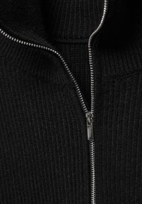 Pull en maille noire avec un col haut et une fermeture éclair argentée. Le tissu a une finition texturée, mettant en valeur les coutures et le détail de la fermeture éclair.
