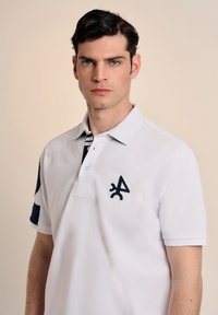 Camisa polo blanca con acentos en azul marino, que presenta un logo en el pecho, cuello con solapa y mangas cortas. Detalle a rayas en el interior del collar.