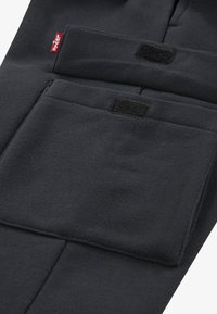 Černé látkové kalhoty s velkou kapsou na suchý zip a menším páskem na suchý zip. Hladká textura s viditelnými švy podél okrajů.