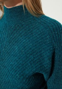 Tyrkysový vlnený rolák se žebrovanou strukturou a mírným leskem, s přiléhavým výstřihem a jemně strukturovanými rameny.