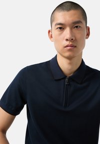 Námořnické polo tričko s límcem na zip, krátkými rukávy a hladkým, texturovaným materiálem s kontrastními tmavými detaily.