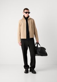 Beige suede jacket, black turtleneck, black trousers, and black shoes. Holding a black tote bag. Sunglasses with a square frame.
