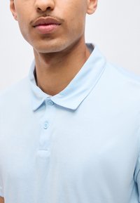 Κοντινό πλάνο ενός ατόμου που φοράει ανοιχτό μπλε πόλο πουκάμισο με γιακά, εμφανίζοντας το λαιμό, τα χείλη και το πάνω μέρος του στήθους του, σε απλό φόντο.