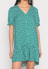 Abito verde da donna con scollo a V, maniche corte con volant e chiusura a bottoni davanti. Il tessuto presenta un motivo floreale bianco.