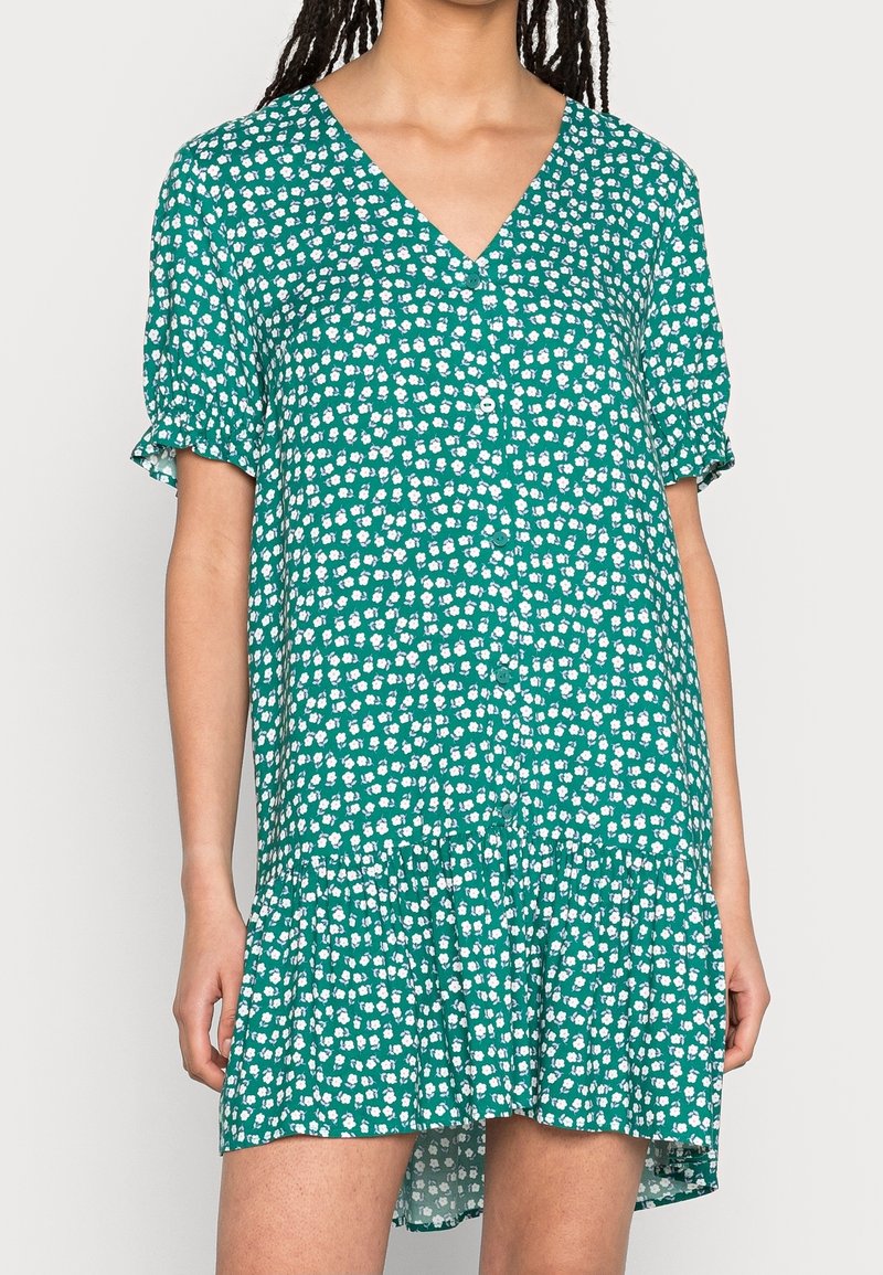 Abito verde da donna con scollo a V, maniche corte con volant e chiusura a bottoni davanti. Il tessuto presenta un motivo floreale bianco.