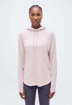 Femme aux cheveux tressés portant un haut à capuche à manches longues rose clair et un legging de sport sombre avec un petit logo sur la cuisse.