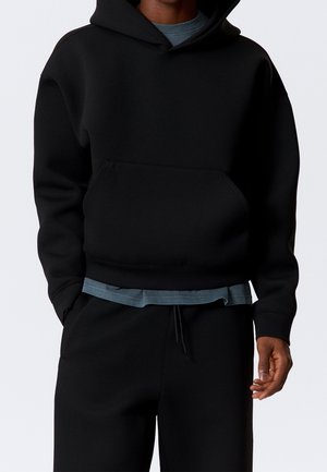 Personne portant un sweat à capuche noir sur un t-shirt gris et un pantalon noir assorti, les mains détendues le long du corps, devant un fond uni.