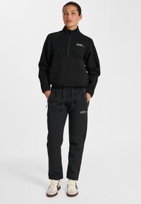 Sweat-shirt de sport noir avec col zippé mi-haut, associé à un pantalon de jogging noir fuselé. Les deux pièces présentent un tissu texturé et de discreet accents de logo.