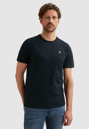 Man met kort bruin haar en baard, die een effen zwarte korte mouwen T-shirt en blauwe spijkerbroek draagt tegen een lichte achtergrond.