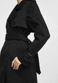 Czarny płaszcz trench z podwójnym zapięciem, szerokim kołnierzem, paskiem w talii oraz teksturowanym detalem z klamrą na mankietach.