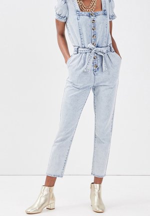 Combinaison en denim bleu clair avec manches courtes, encolure carrée, boutons sur le devant, ceinture à la taille et poches latérales. Associée à des bottines dorées.