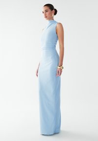 Světle modré šaty bez rukávů s vysokým límcem, přiléhavým živůtkem a dlouhou sukní. Texturovaná látka a jemné boční řasení. Viditelný zlatý náramek na zápěstí.