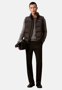 Czarna pikowana kamizelka z wysokim kołnierzem na szarym swetrze, do tego czarne spodnie i wypolerowane czarne buty, trzymająca czarną kurtkę.