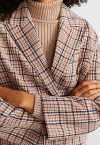 Karo-Blazer in Beige, Pink und Navy mit Reverskragen. Getragen über einem gerippten beigen Rollkragenpullover. Besitzt einen lässigen Schnitt und umgeschlagene Manschetten.