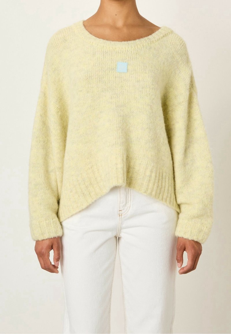 Personne portant un pull duveteux jaune clair à col rond et ourlet asymétrique, associé à un pantalon blanc.