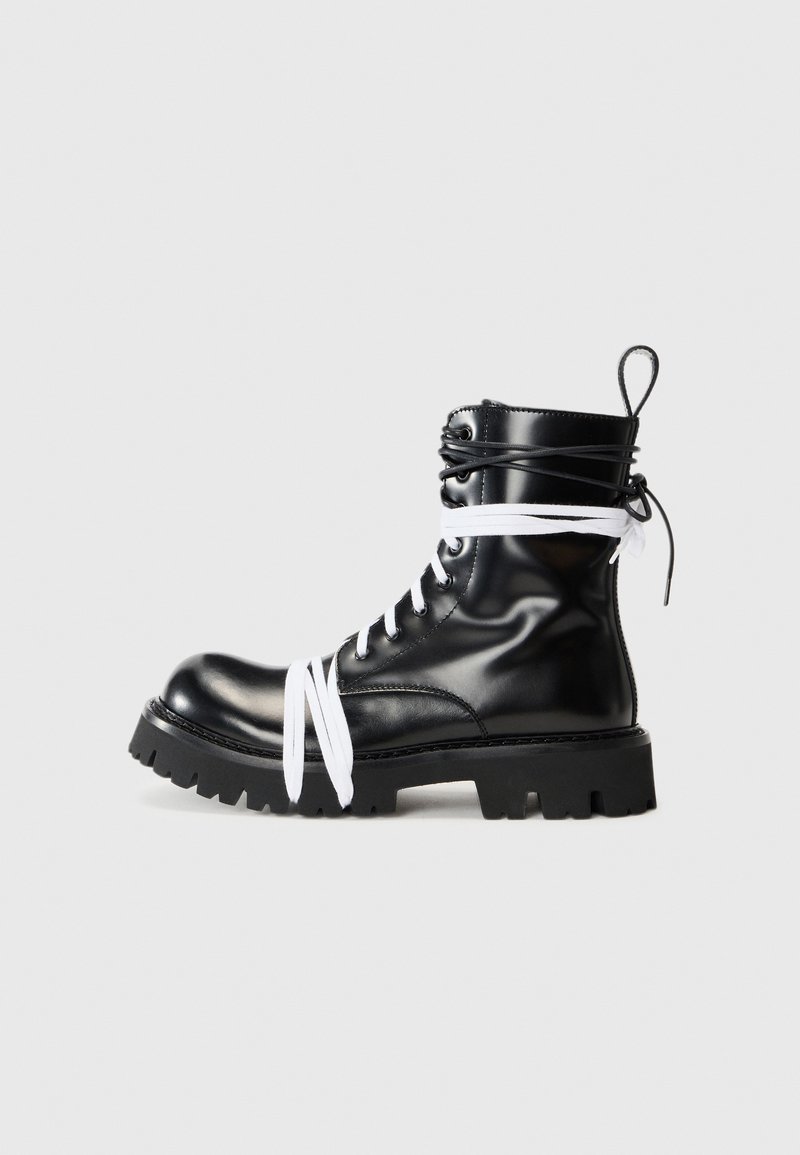 Bota de combate de cuero negro con cordones blancos, punta redonda, suela negra resistente y detalle de costura. Cuenta con un lazo en la parte posterior para mayor comodidad.