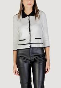 Cardigan bianco con bordi neri, chiusura a bottoni e colletto arrotondato. Presenta due tasche nere decorative. Abbinato a pantaloni di pelle neri.