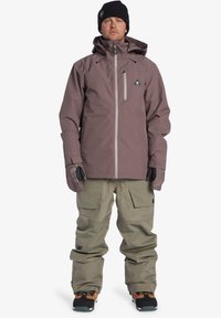 Heren sneeuwjas in zacht grijs met een ritszak op de borst, gecombineerd met losse beige sneeuwbroeken. Zwarte handschoenen en een beanie maken de outfit compleet.