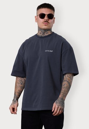 T-shirt surdimensionnée gris foncé avec des manches courtes et un col rond, arborant un logo blanc sur la zone gauche de la poitrine. Texture en coton doux.